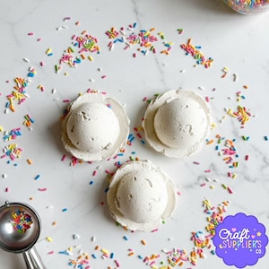 Pode incluir: Três objetos brancos, texturizados, em forma de sorvete estão em uma superfície branca, cercados por granulado colorido. Uma colher de sorvete prateada com granulado está à esquerda. Os objetos estão em um prato em forma de flor. O logotipo da Craft Supplier's Co. está no canto inferior direito.