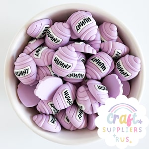 May include: A white bowl filled with lavender-colored resin beads shaped like honey pots. Each bead has the word "HUUNY" in black lettering. The beads are designed for crafting and are a light purple color.