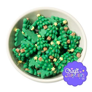 May include: A white bowl filled with numerous small, green, flat Christmas tree-shaped embellishments. Each tree features a light yellow star at the top, brown trunk, and tiny colorful dots representing ornaments. A purple logo reads Craft Suppliers Co.
