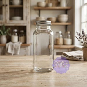 Op de afbeelding: Een heldere, rechthoekige glazen pot met een zilveren deksel. De pot is leeg en staat op een houten oppervlak. De achtergrond omvat een keuken met planken, kommen en een lavendelplant. De pot heeft de tekst "Craft Suppliers Co."