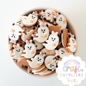 May include: A bowl filled with Halloween-themed cookies. The cookies are shaped like ghosts with pink bows, orange pumpkins, and black eyes. The cookies are white with brown edges and are in a white bowl.