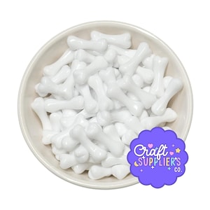 May include: A round white bowl filled with numerous small, smooth, white bone-shaped beads. A purple scalloped sticker with the text Craft SUPPLIER'S CO. is positioned in the lower right corner of the image, partially overlapping the bowl.
