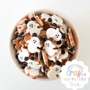Puede incluir: Un cuenco de cerámica blanco lleno de adornos artesanales con temática de Halloween. Los adornos presentan formas de fantasmas blancos con orejas y ojos negros, y mejillas rosas. Los fantasmas están rodeados de formas marrones y beige.