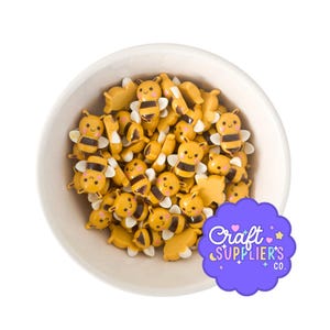 May include: A white bowl filled with small, yellow and black bee-shaped cabochons featuring smiling faces and white wings. A purple scalloped sticker in the bottom right corner displays the text Craft SUPPLIER'S CO. with small star and heart icons.