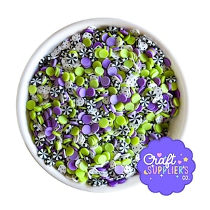 Puede incluir: Un cuenco blanco lleno de chispas de arcilla polimérica de colores. Las chispas son de varias formas y colores, incluyendo verde, morado, negro y blanco. El texto "Craft Supplier's Co." está sobre un fondo morado en la esquina inferior derecha.