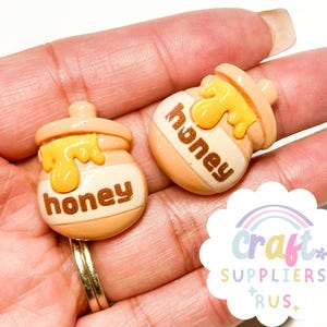 Może przedstawiać: Dwa charmsy w kształcie słoika miodu z jasnopomarańczową podstawą, żółtym kroplami miodu i brązowym napisem "honey". Charmsy są małe i prawdopodobnie przeznaczone do rękodzieła lub tworzenia biżuterii. Tło to dłoń.