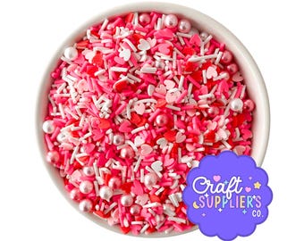 Pode incluir: Uma tigela branca cheia com uma mistura de granulados vermelhos, rosas e brancos, incluindo peças em forma de coração, bastões finos e esferas redondas peroladas. Uma etiqueta roxa recortada no canto apresenta o texto Craft SUPPLIER'S CO. em branco.