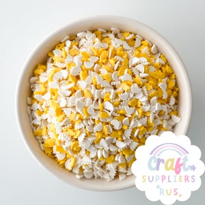 Könnte beinhalten: Eine Schüssel gefüllt mit weißen und gelben Streuseln in Form von Halbmonden und kleinen Blüten. Die Streusel eignen sich perfekt zum Dekorieren von Kuchen, Cupcakes und Keksen.