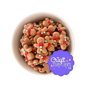 Puede incluir: Un cuenco blanco lleno de decoraciones con forma de hombre de jengibre marrón. Cada hombre de jengibre tiene una cara sonriente, una pajarita roja y detalles azules y blancos. Se ve un logotipo morado con texto.