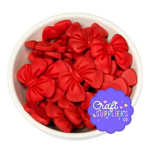 May include: A white bowl filled with numerous small, matte red bow-shaped embellishments. A purple scalloped sticker with the white text Craft Suppliers Co. is positioned over the lower right portion of the bowl containing the red bows.
