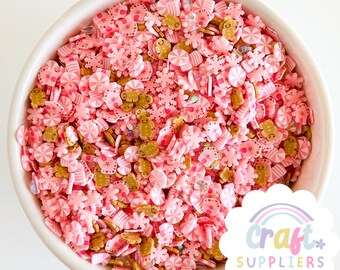 Pode incluir: Uma tigela branca cheia de granulados de argila polimérica rosa e dourada. Os granulados têm várias formas, incluindo bonecos de gengibre, flocos de neve e bengalas doces. Os granulados são para artesanato e estão em um fundo branco.