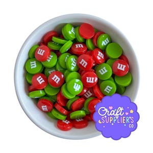 May include: A white bowl filled with red and green disc-shaped embellishments. Each disc has a white "m" printed on it. The embellishments are scattered throughout the bowl, creating a colorful and festive appearance.