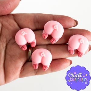 以下が含まれることがあります： 赤い蹄を持つ4つのピンク色の豚のお尻チャームが表示されています。チャームは手の中にあり、指には指輪が付いています。「Craft Suppliers Co.」の紫と白のロゴが右下にあります。