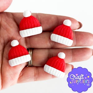 Puede incluir: Cuatro mini gorros de Papá Noel. Cada gorro es rojo con un borde blanco y un pompón blanco. Los gorros son pequeños, probablemente para manualidades o decoración. Se sostienen en una mano.