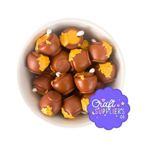 Può includere: Una ciotola bianca riempita con numerosi piccoli ciondoli marroni a forma di vasetto, ognuno caratterizzato da un design a goccia di miele giallo e un manico di cucchiaio bianco. Un adesivo viola smerlato con il testo Craft SUPPLIER'S CO. è in basso a destra.