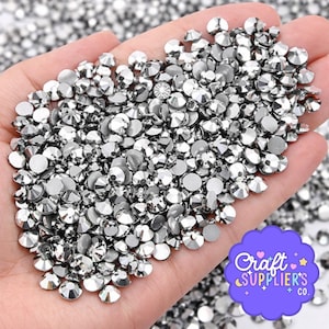 May include: A handful of silver rhinestones, each with multiple facets, reflecting light. The gemstones are round and scattered, creating a sparkling effect. The image includes a logo that says "Craft Supplier's Co."