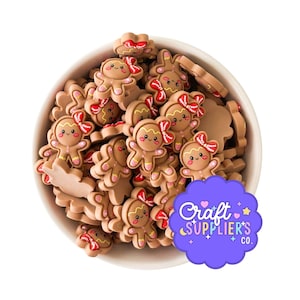 May include: A white bowl overflowing with brown gingerbread man-shaped craft embellishments. Each gingerbread man has a red bow, pink accents, and a smiling face. A purple sticker with the text "Craft SUPPLIER'S CO." is in the bottom right corner.