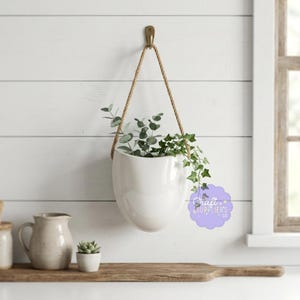 Może przedstawiać: Biały ceramiczny kubek z czarnym uchwytem i napisem "But First, Coffee" czarną, pogrubioną czcionką. Kubek stoi na białej powierzchni.