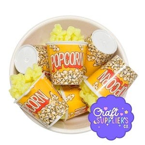Puede incluir: Una colección de mini vasos de palomitas de maíz llenos de palomitas amarillas, dispuestos en un cuenco blanco. Los vasos son amarillos con "POP CORN" impreso en rojo. Un logotipo púrpura con texto está en la esquina inferior derecha.