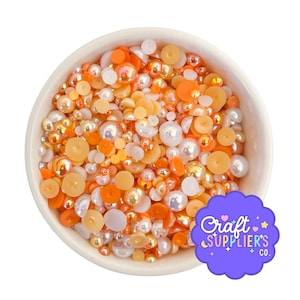 May include: A white bowl filled with a mix of orange, yellow, and white beads. The beads vary in size and shape, including round, flat, and pearl-like. The bowl has a purple sticker with the text "Craft Supplier's Co."