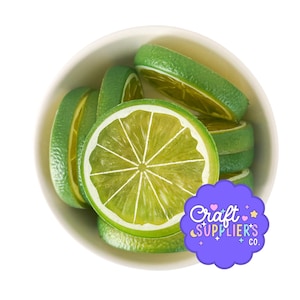 May include: A white bowl filled with several artificial lime slices. The slices feature a realistic green rind and a translucent, light green interior with white segment lines. A purple scalloped sticker with the text Craft SUPPLIER'S CO. is visible.
