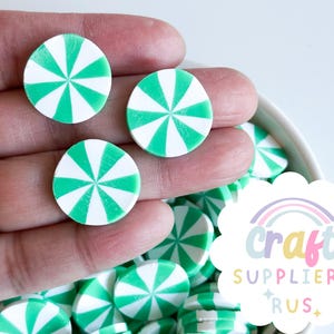 May include: A close-up of a hand holding green and white peppermint candy-shaped polymer clay embellishments. The candy is in a bowl with other candy pieces.