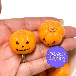 May include: Two bright orange pumpkin-shaped charms with carved jack-o'-lantern faces featuring triangular eyes and jagged mouths. One charm is attached to a silver-toned metal ring base. A purple scalloped logo reads Craft SUPPLIER'S CO.