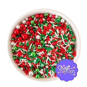 May include: A white bowl filled with a festive mix of red, green, and white sprinkles. The sprinkles include small red and green spheres, white and green rods, and small white pearl-like sprinkles. The bowl is set against a white background.