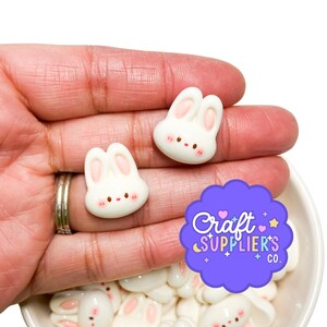 May include: Two small white rabbit-shaped cabochons with pink inner ears and cheeks, brown eyes, and a brown nose. A purple scalloped logo with the text Craft SUPPLIER'S CO. is overlaid on the image, which shows a bowl filled with similar pieces.