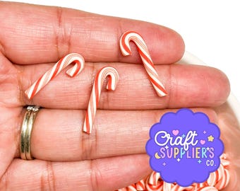 May include: Three small, curved candy cane ornaments featuring a classic red and white striped pattern. The candy canes are smooth and glossy, with a distinct hook shape at the top. A purple circular logo with the text Craft SUPPLIER'S CO. is visible.