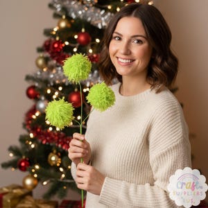 Puede incluir: Una mujer sostiene un tallo de tres flores de pompón verde brillante. Las flores son esponjosas y redondas, con una apariencia texturizada. El fondo incluye un árbol de Navidad decorado con adornos rojos y dorados.