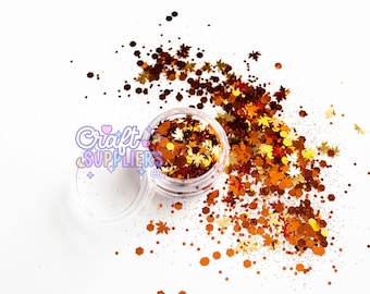Könnte beinhalten: Ein durchsichtiges Plastikglas gefüllt mit herbstlichen Glitzer, darunter Blattformen und sechseckige Teile in Braun-, Orange- und Goldtönen. Der Glitzer ist auf einer weißen Oberfläche verstreut, was für eine festliche Stimmung sorgt.