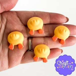 以下が含まれることがあります： オレンジ色の足が付いた、黄色のアヒルの尻のチャームが4つ表示されています。チャームは丸みを帯びた形状で、表面はテクスチャ加工されています。チャームはおそらく、工芸品やジュエリー作りに使用されます。