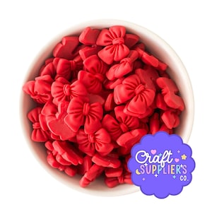May include: A white bowl filled with numerous small, bright red bow-shaped decorations. The bows are densely packed, creating a textured surface. A purple sticker with the text "Craft Supplier's Co." is in the bottom right corner.