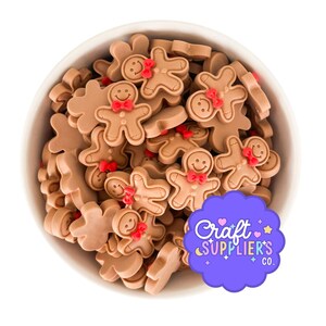 Puede incluir: Un cuenco blanco lleno de adornos artesanales con forma de hombre de jengibre marrón. Cada hombre de jengibre tiene una pajarita roja y una cara sonriente. Un logotipo púrpura con el texto "Craft SUPPLIER'S CO." está en la esquina inferior derecha.