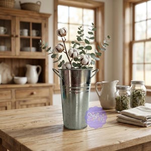 Puede incluir: Un cubo de metal galvanizado con asas contiene tallos de algodón y ramas de eucalipto. El cubo está sobre una mesa de madera, con una jarra blanca y frascos de vidrio en el fondo. La imagen tiene una estética rústica.