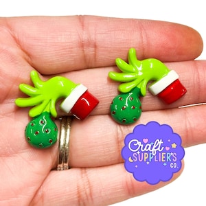 May include: Two flat-backed charms shaped like bright green furry hands wearing red and white cuffs, each holding a green ornament with small red dots. The charms feature a glossy finish and are designed for crafting or decorative applications.