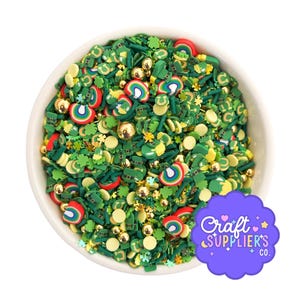 May include: A white bowl filled with St. Patrick's Day-themed sprinkles. The sprinkles include green shamrocks, leprechaun hats, rainbows, and gold beads. The bowl is set against a white background. A purple logo with the text "Craft Supplier's Co." is in the bottom right corner.