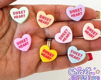 May include: A collection of heart-shaped charms in pastel colors, each inscribed with the words "SWEET HEART" in red. The charms are in shades of pink, yellow, blue, and lavender. The image also includes the logo for "Craft Suppliers Co."