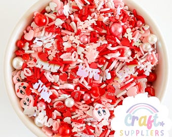 Pode incluir: Uma tigela branca cheia de uma mistura de granulados vermelhos, rosa e brancos. Os granulados incluem corações, lábios e a palavra "LOVE". Há também contas peroladas e outras formas decorativas. O tema geral é o Dia dos Namorados.