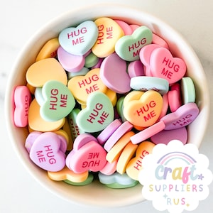 Pode incluir: Uma tigela branca cheia de doces coloridos em forma de coração. Cada doce tem as palavras "Hug Me" impressas em vermelho.