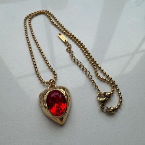 May include: A gold-colored necklace with a heart-shaped pendant. The pendant features a large, oval, red gemstone set in a textured gold frame. The chain is a delicate ball chain with a lobster clasp.