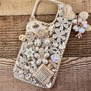 May include: A clear phone case decorated with rhinestones, pearls, and charms. The charms include a white carriage, a gold heart with the text "I love you", and a white flower.