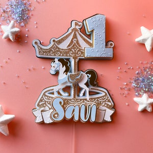 May include: A white and gold carousel cake topper with a blue horse and the number one. The topper also includes the name "Savi" in blue lettering.