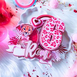 May include: A pink and white birthday cake topper with a cheetah design, the number 6, and the name "Remi". The topper includes a cheetah illustration, hearts, and decorative elements. The background is a fluffy white surface.