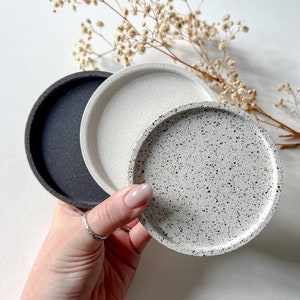 May include: Three round coasters, one black, one white, and one white with black speckles. The coasters are made of a textured material and are stacked on top of each other.
