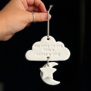 May include: White ceramic cloud ornament with the text "Welcome to the world Harry & Evie" hanging from a grey string. The cloud is attached to a white ceramic star and crescent moon.