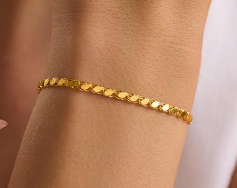 May include: A thin, yellow gold bracelet featuring a delicate, repeating textured link design that wraps around the wrist. The metal has a polished, reflective finish that catches the light, creating a subtle shimmer along the entire length of the chain.