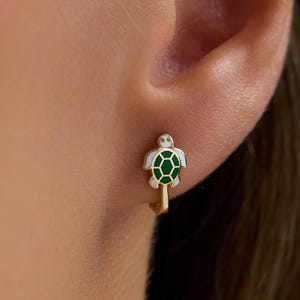 May include: A gold hoop earring featuring a small turtle charm. The turtle has a green enamel shell with a white border and a gold head and limbs. The earring is secured through the earlobe, showcasing the detailed turtle design in a compact size.