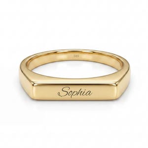 May include: A polished gold band ring featuring a flat, rectangular front surface engraved with the name Sophia in a cursive script. The ring has a smooth, reflective finish and a consistent metallic gold tone throughout the entire piece.